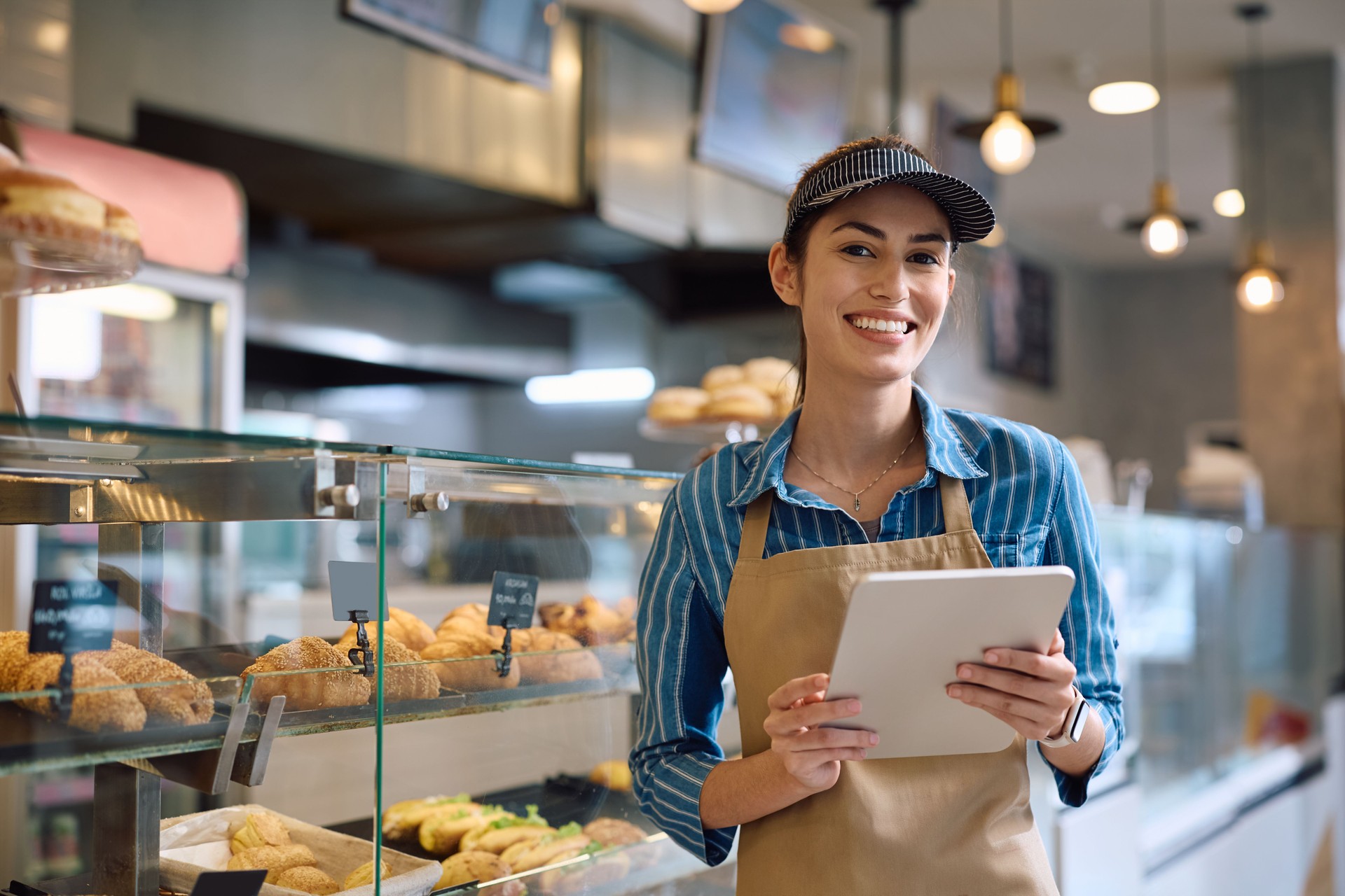 Young happy woman working on digital tablet in a bakery and looking at camera.
